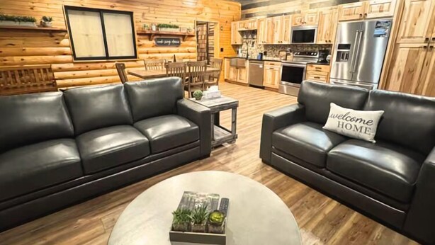 A log cabin interior with a living area featuring a black leather sofa, a coffee table with a plant, and a kitchen with wooden cabinets and stainless steel appliances. - NEW! Little Ranch Therapy - Hot Tub, Fireplace (Traverse City)