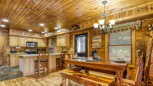 A wooden cabin interior with a kitchen, dining area, and a large window with blinds. - Elk Crossing 3 Bedroom Cabin (Pigeon Forge)