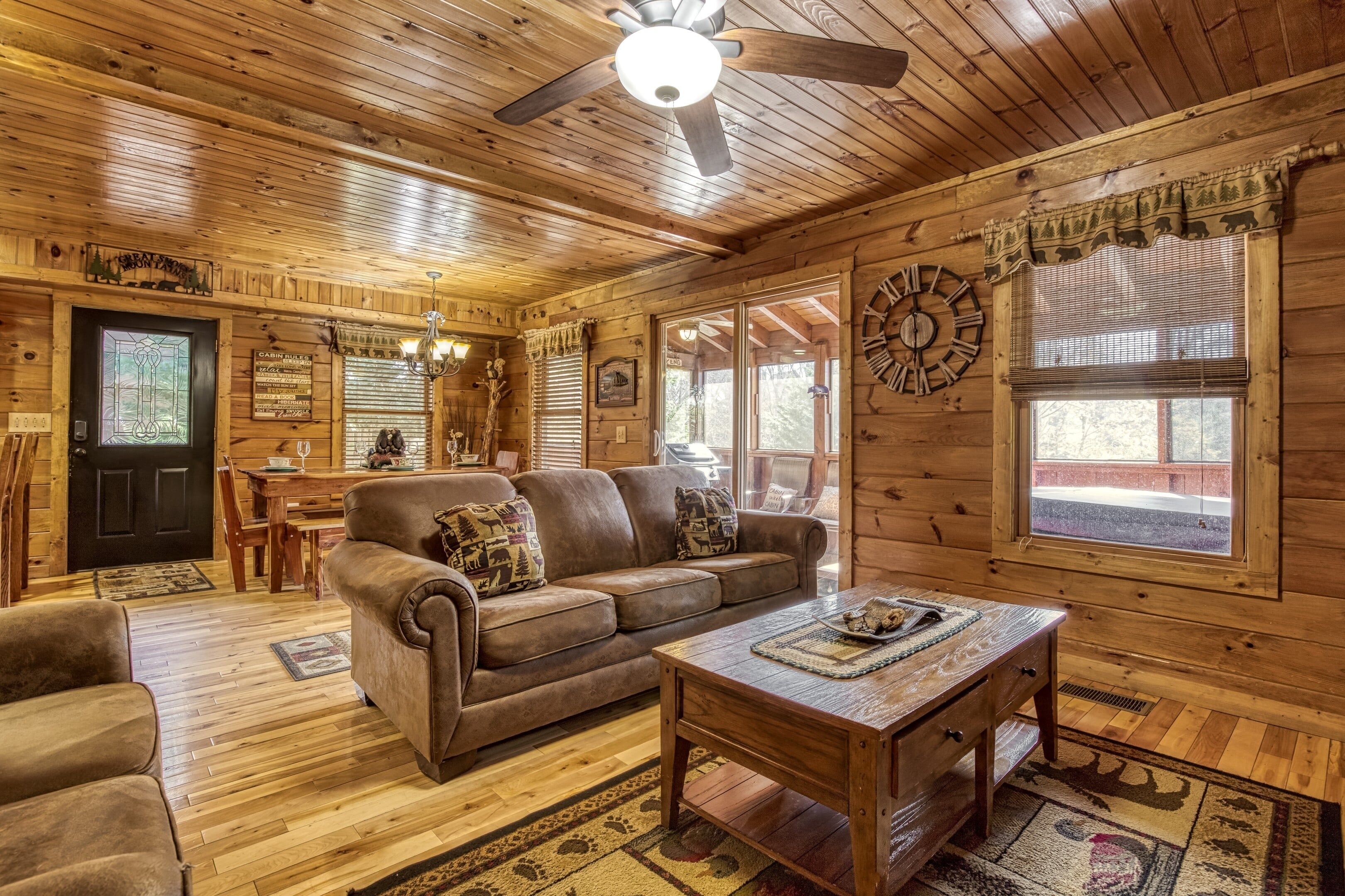 A log cabin interior with a wooden ceiling, a ceiling fan, a dining area with a table and chairs, a brown leather sofa, and a wooden coffee table.