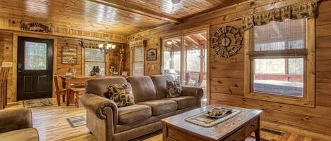 A log cabin interior with a wooden ceiling, a ceiling fan, a dining area with a table and chairs, a brown leather sofa, and a wooden coffee table.