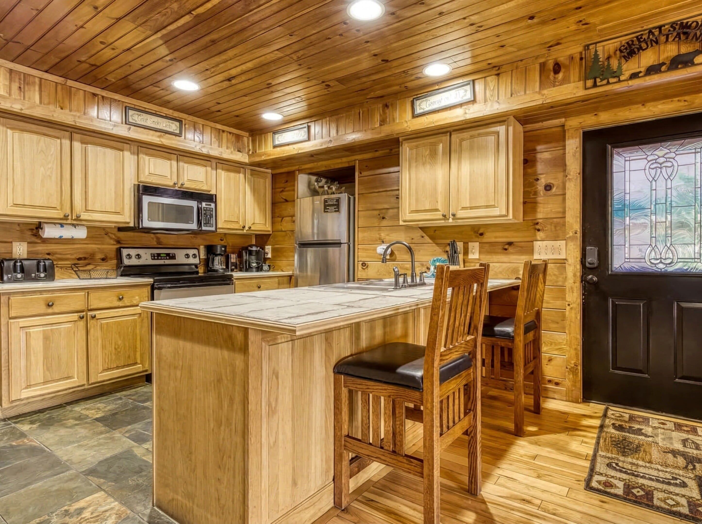 Interior de una cabaña de madera con cocina, que incluye una isla central, armarios de madera, un refrigerador y una puerta negra con panel de vidrio.