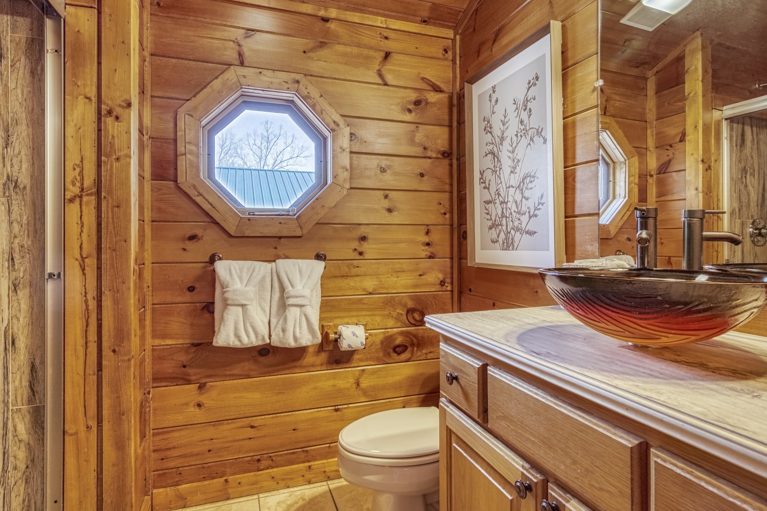 Baño de cabaña de madera con tragaluz hexagonal, inodoro blanco, tocador de madera con lavabo de vidrio y una impresión botánica enmarcada en la pared.