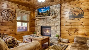 A cabin interior with a stone fireplace, wooden walls, a ceiling fan, a clock, and a welcome sign. - Elk Crossing 3 Bedroom Cabin (Pigeon Forge)