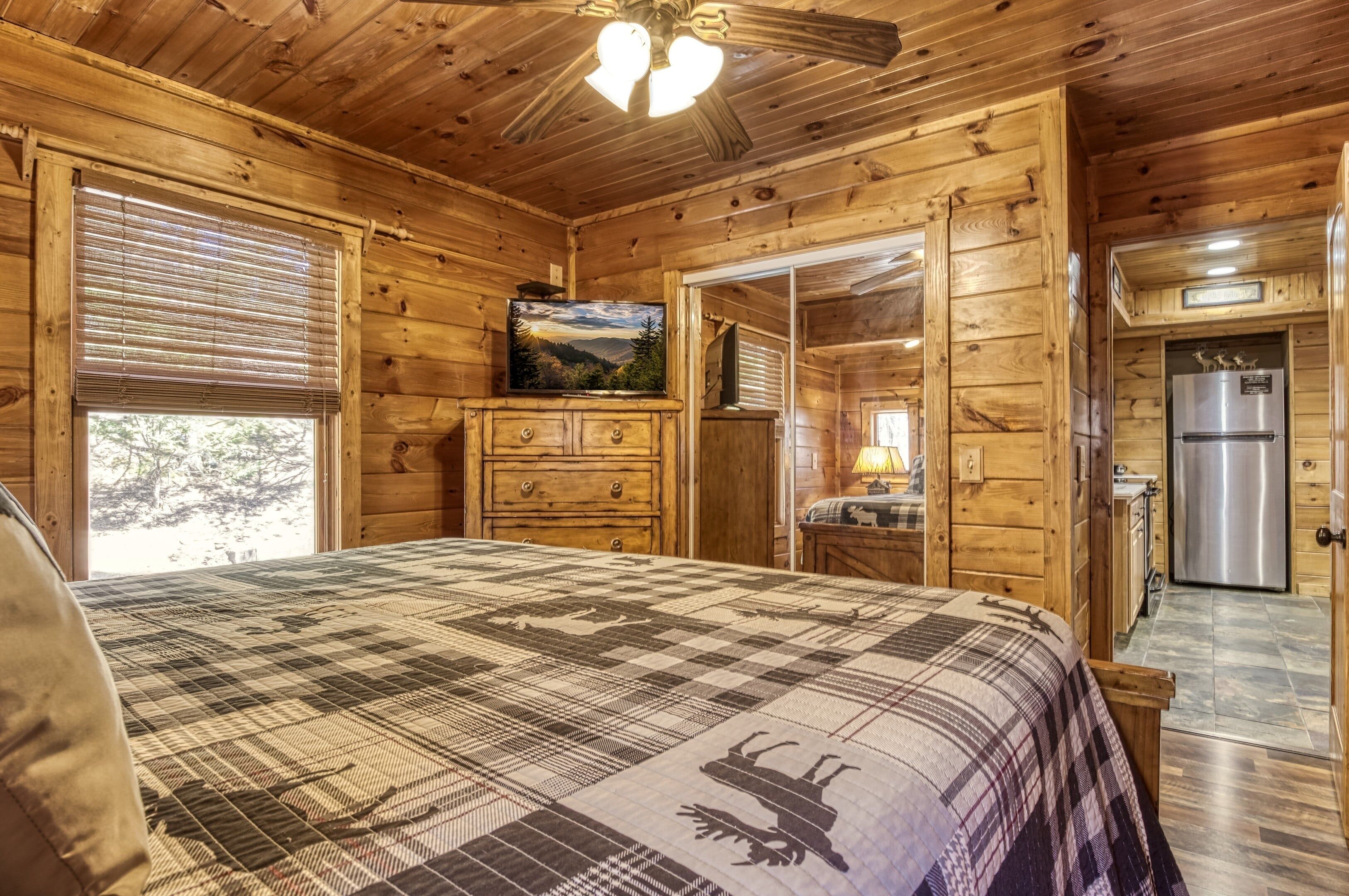 A wooden cabin bedroom with a bed, dresser, television, and a refrigerator.