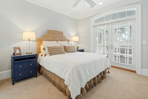 Une chambre à coucher avec un grand lit, deux tables de chevet avec des lampes, une tête de lit en bois et une vue à travers un ensemble de volets blancs.