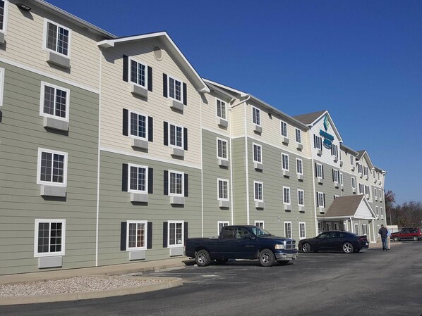A multi-story hotel with a row of rooms on each floor, a parking lot with several vehicles, and a clear blue sky. - Value Place Louisville Ky South (Fairdale)