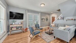 Un salon avec un canapé gris, des fauteuils bleus, une table basse en bois et un téléviseur fixé au mur.