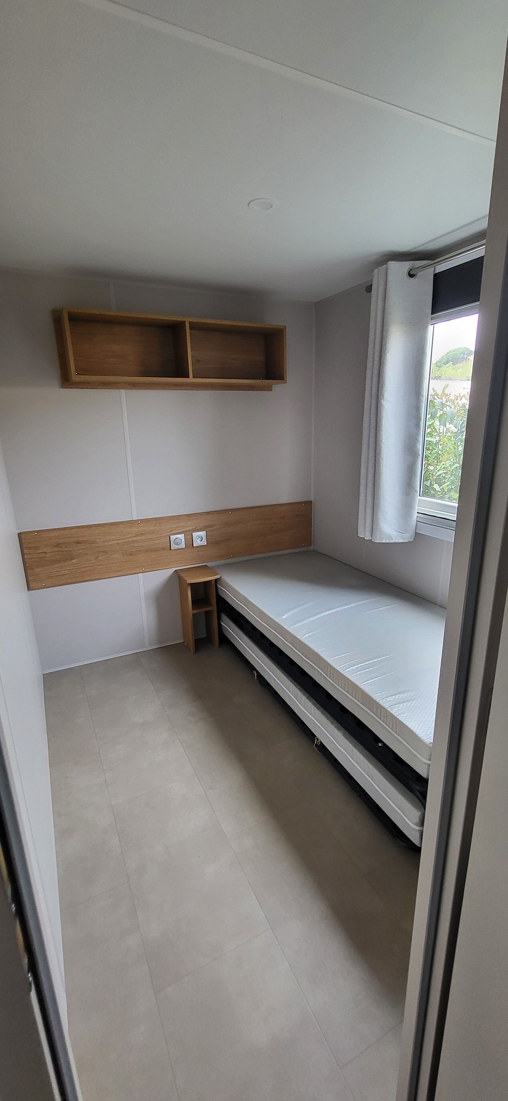 A small, compact room with a single bed, a wooden shelf above the bed, a small stool, and a window with curtains.