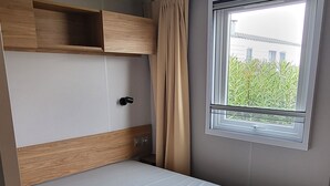 Une pièce de taille modeste, avec un lit simple, une petite fenêtre avec des rideaux et une étagère fixée au mur.