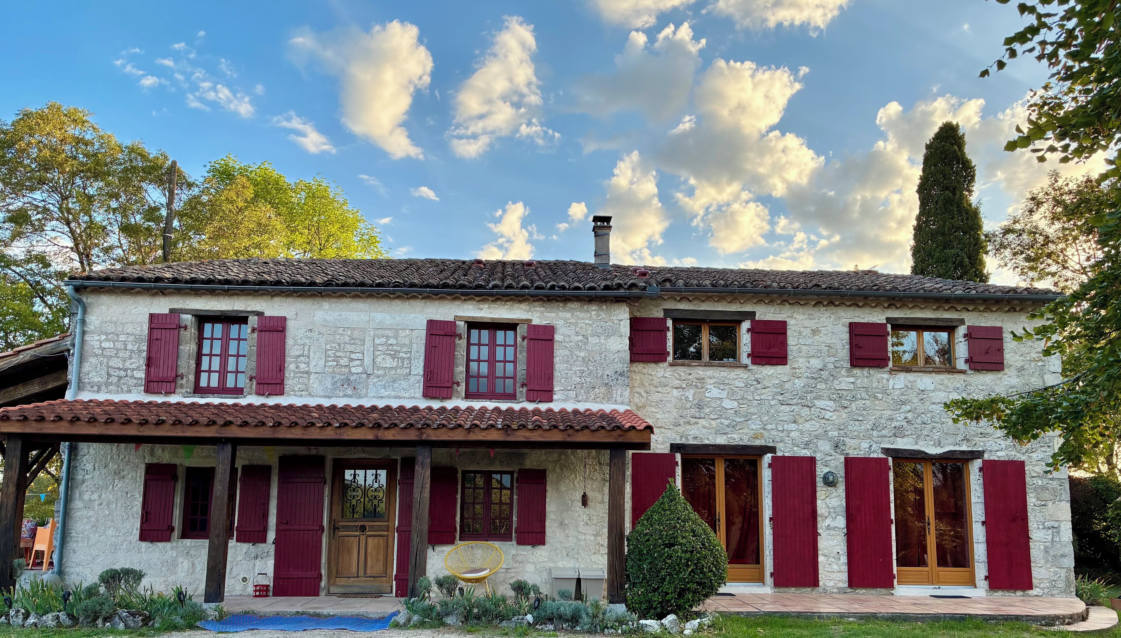 A two-story stone house with red shutters, a tiled roof, and a small porch with a chair.