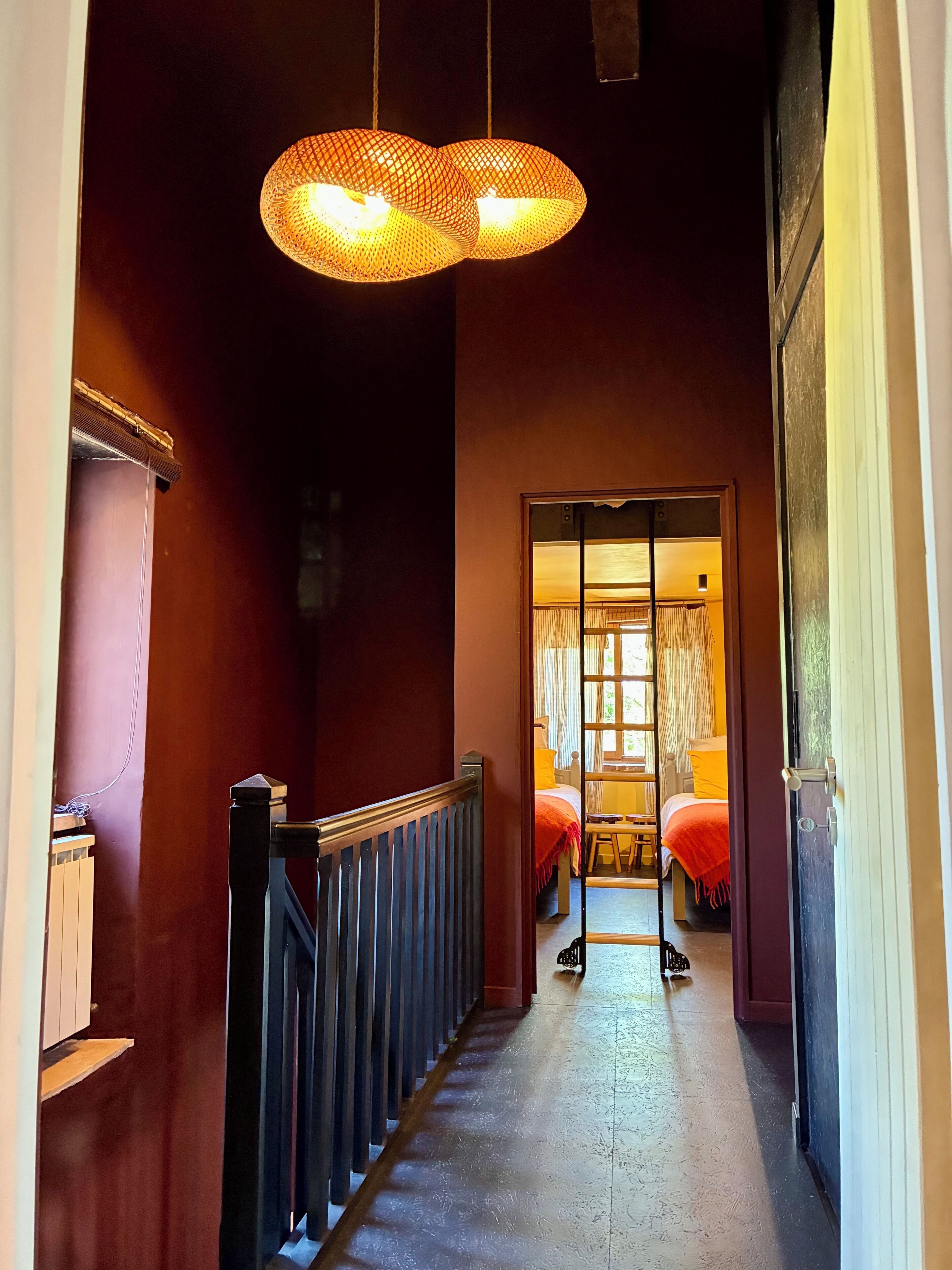 A hallway with a wooden railing, a bed with a red blanket, and a hanging light fixture.
