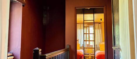 A hallway with a wooden railing, a bed with a red blanket, and a hanging light fixture.
