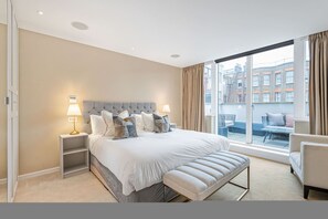 A modern hotel room with a large bed, bedside lamps, a bench, and a view of the city through large windows. - Luxury Kensington Central AC Private Terrace (London)