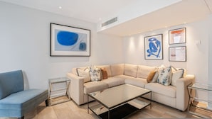 Ein modernes Wohnzimmer mit einem beigefarbenen Ecksofa, einem Glastisch und einem blauen Sessel. An den Wänden befinden sich gerahmte Kunstwerke und ein kleiner Beistelltisch.