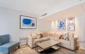 A modern living room with a beige sectional sofa, a glass coffee table, and a blue armchair. There are framed artworks on the walls and a small side table. - Luxury Kensington Central AC Private Terrace (London)