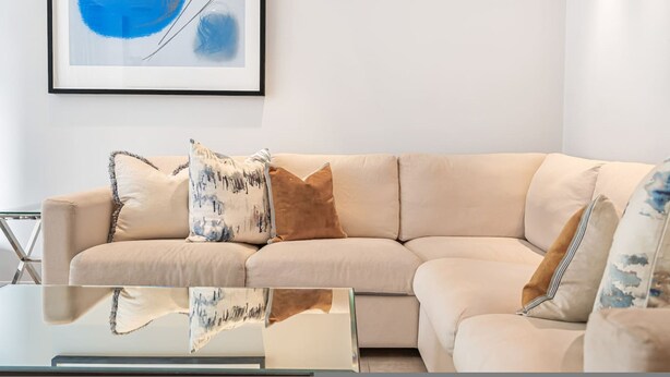 Ein beiges Modulsofa mit gemusterten Kissen, ein Glastisch und ein gerahmtes abstraktes Gemälde an der Wand.