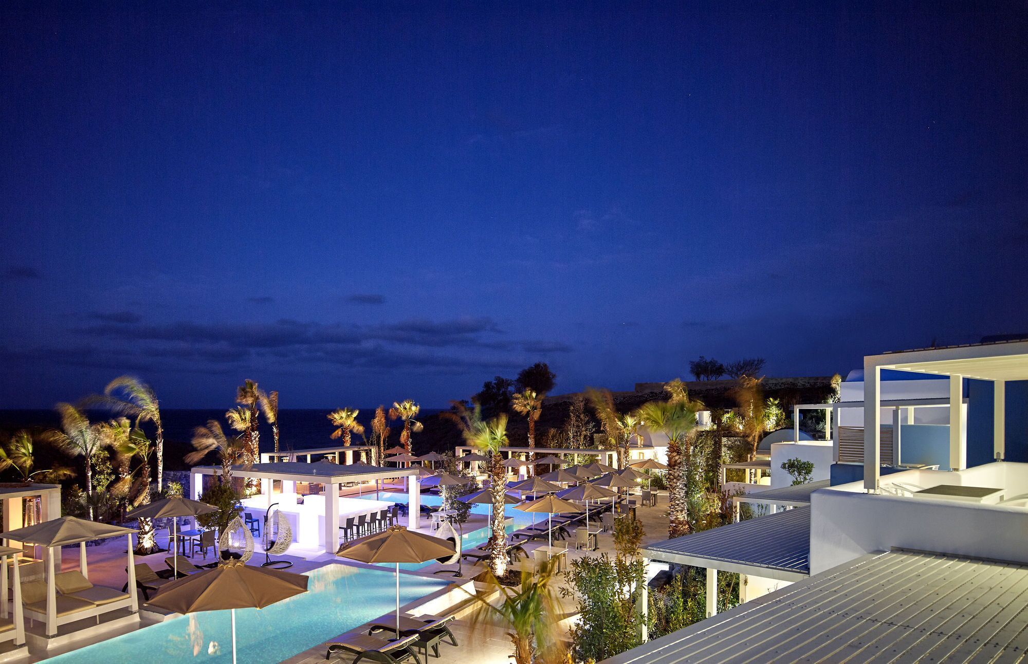Zona de piscina en un complejo turístico con sillones de playa, sombrillas y un bar, de noche.