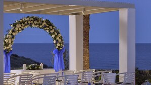 An outdoor wedding setup with white chairs and tables, a floral arch, and lanterns. - Aurora Luxury Hotel & Spa (Santorini)