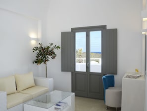 A modern living room with a white sofa, a glass coffee table, a blue chair, and a view of the sea through a grey door. - Aurora Luxury Hotel & Spa (Santorini)