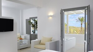 A modern hotel room with a bed, a seating area with a cushioned bench, a small table with a bowl of fruit, a flat-screen TV mounted on the wall, and a large window offering a view of the outdoors. - Aurora Luxury Hotel & Spa (Santorini)