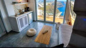 A modern kitchen with a large window overlooking a scenic view, a wooden table, and a cozy seating area. - Nefeli Homes (Santorini)