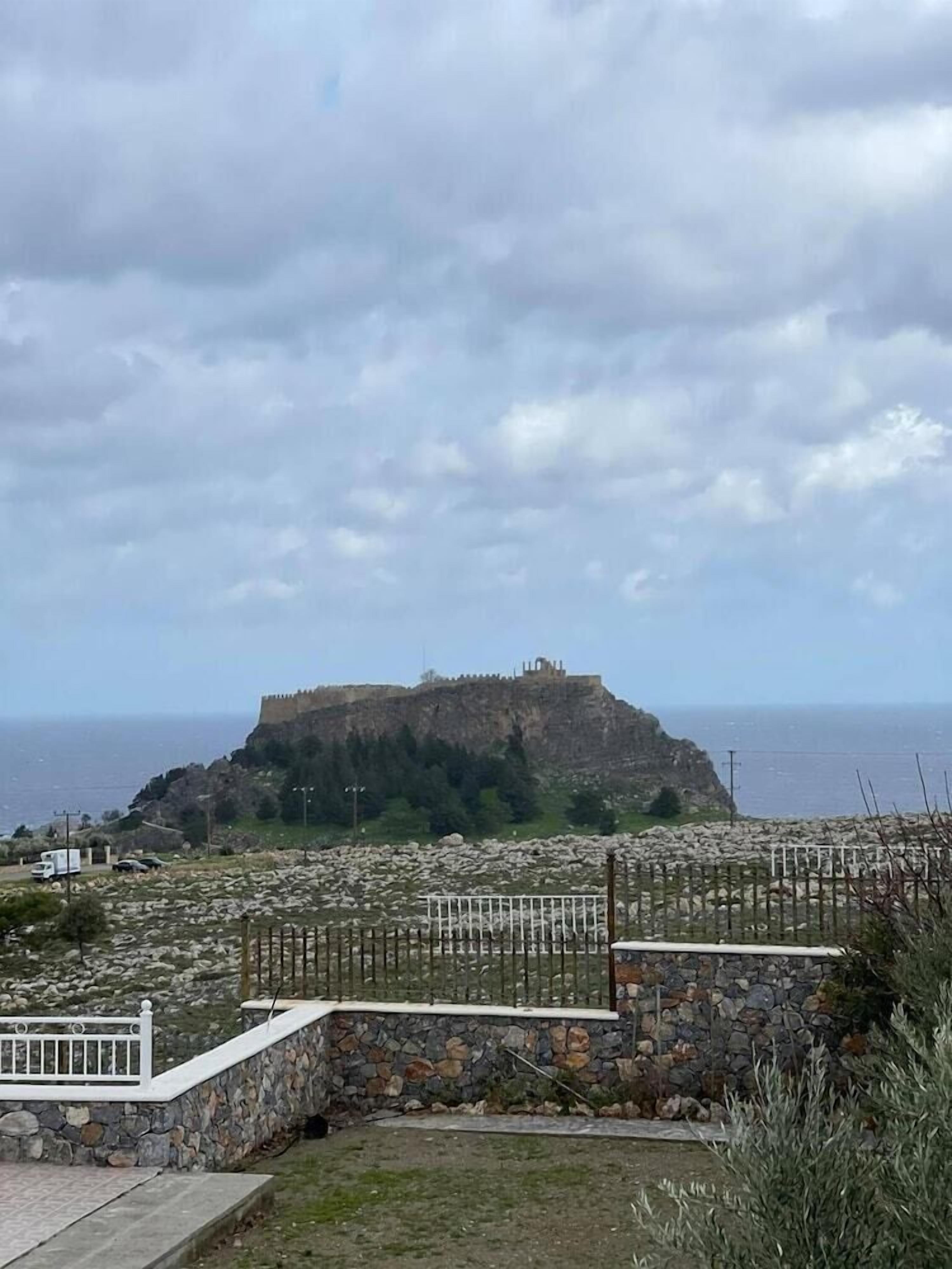 A coastal view with a historic fort on a hill, a stone wall, and a fenced area.