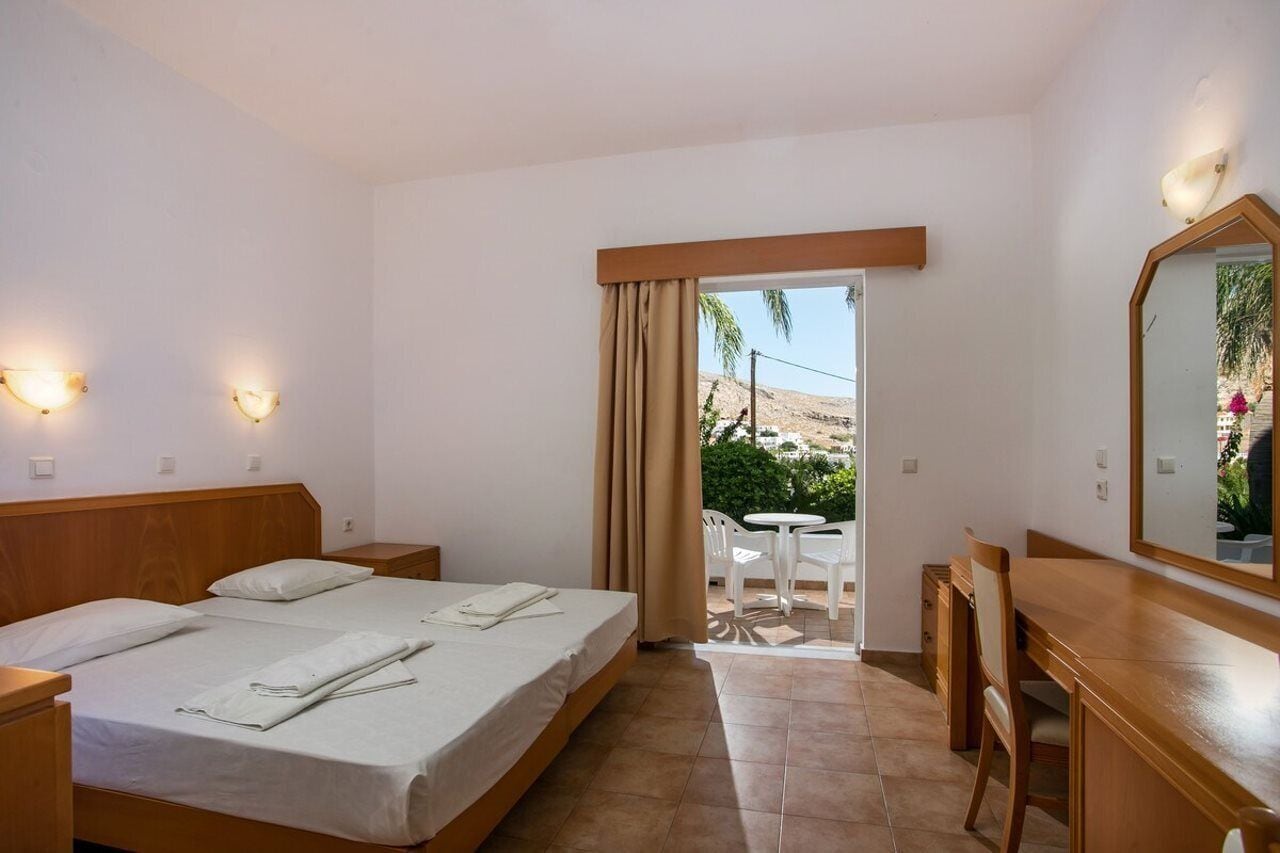 Habitación de hotel con cama, mesitas de noche, escritorio, silla y espejo. Se ve una ventana con cortinas y vistas a zonas verdes.