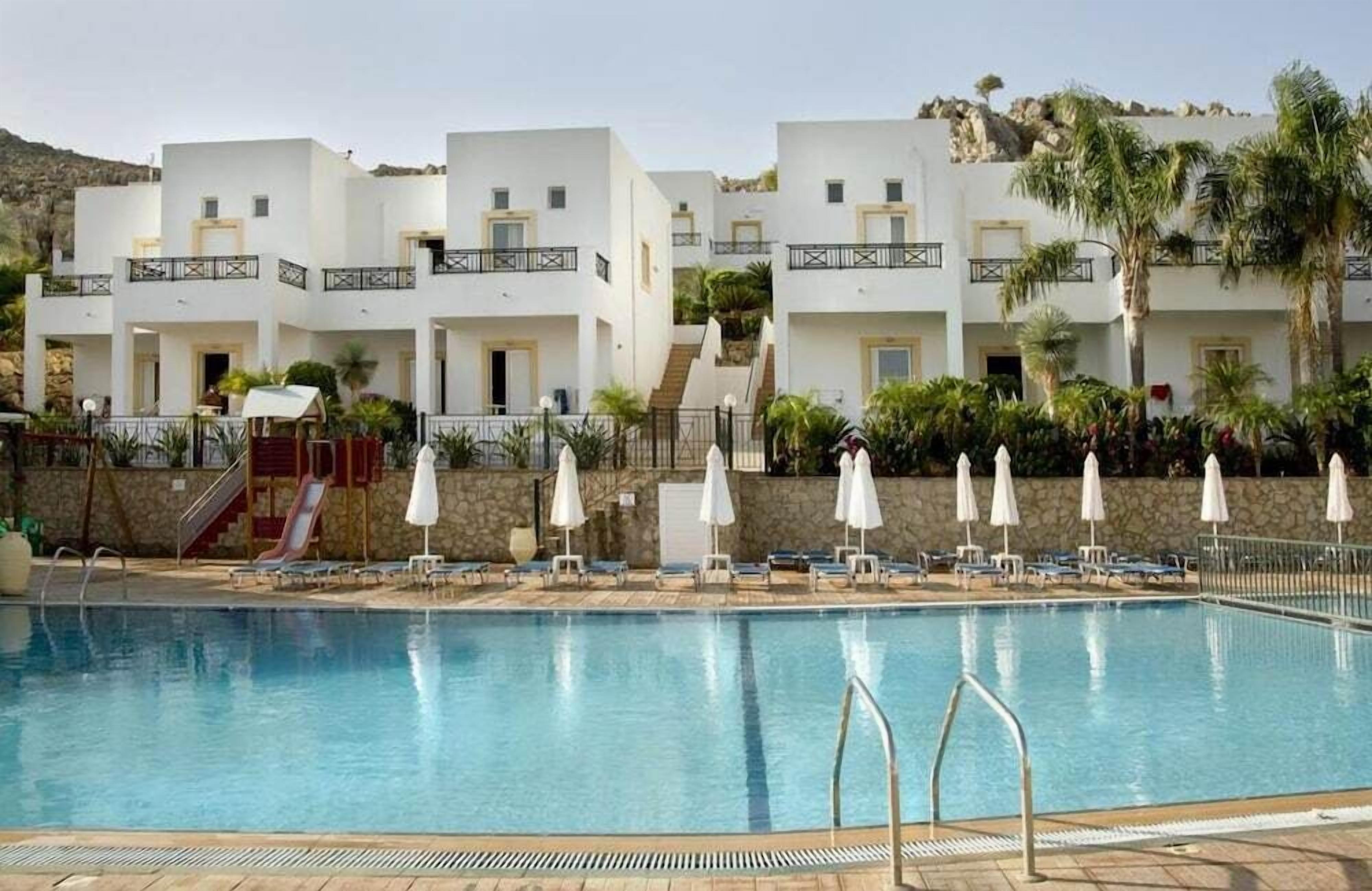 En pool med liggestole og parasoller, omgivet af hvide bygninger med altaner.