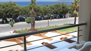 Een balkon met witte kunststof stoelen en een tafel, met uitzicht op een parkeerplaats met auto’s en bomen, en een vergezicht op zee en heuvels in de verte.