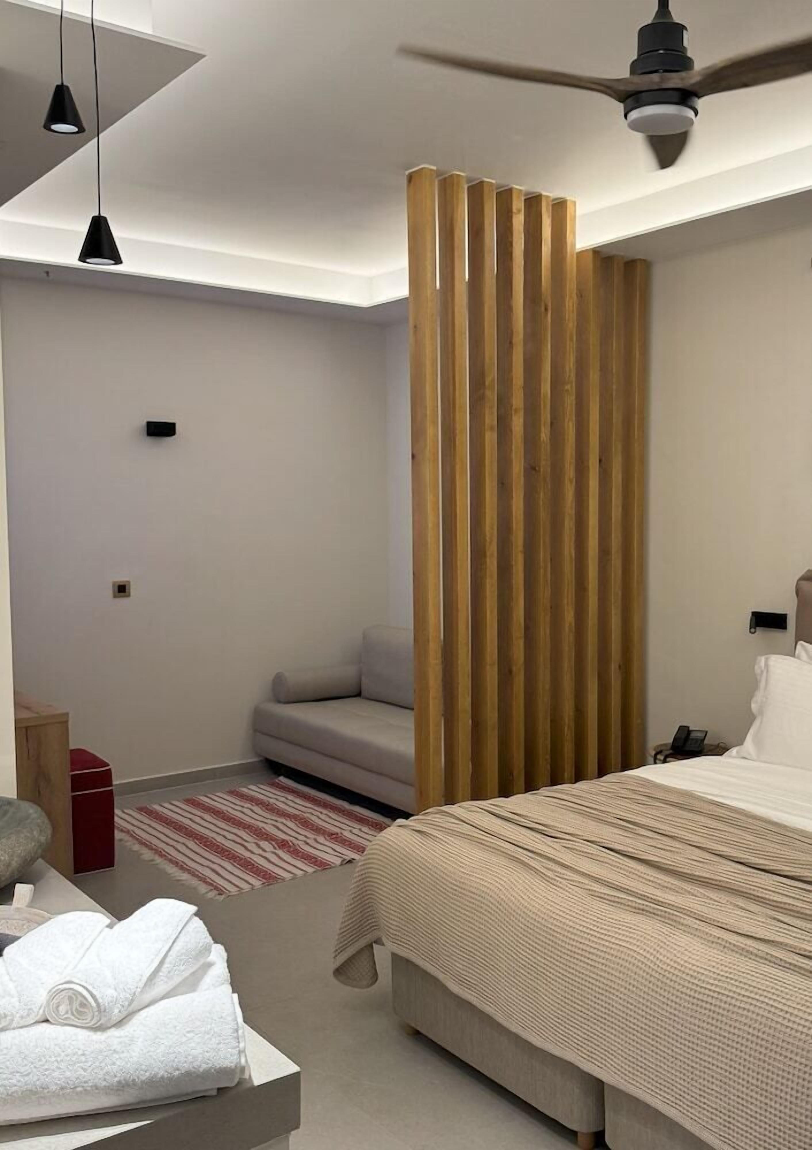 A modern hotel room with a bed, a sofa, a wooden partition, a ceiling fan, and a red and white striped rug.