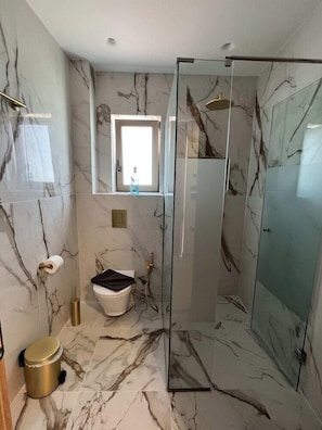 A modern bathroom with a glass shower enclosure, marble walls, and a toilet with a black lid. - The Noverian Antama Organic Beach Resort Chania (Chania)