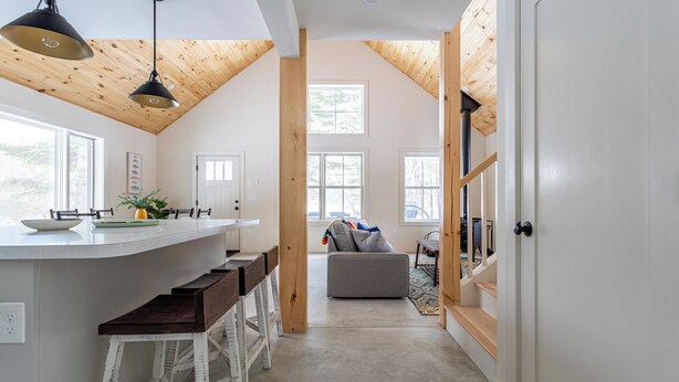 A kitchen with a central island, wooden beams on the ceiling, and a view to a living area with a sofa and windows. - Lakefront with Private Dock & Canoe Kenoza Den (Jeffersonville)