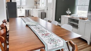 A modern kitchen with white cabinets, a wooden dining table with a floral runner, and a view of the outside through a large window. - Kenoza House with Private Lake and Kayaks (Callicoon)