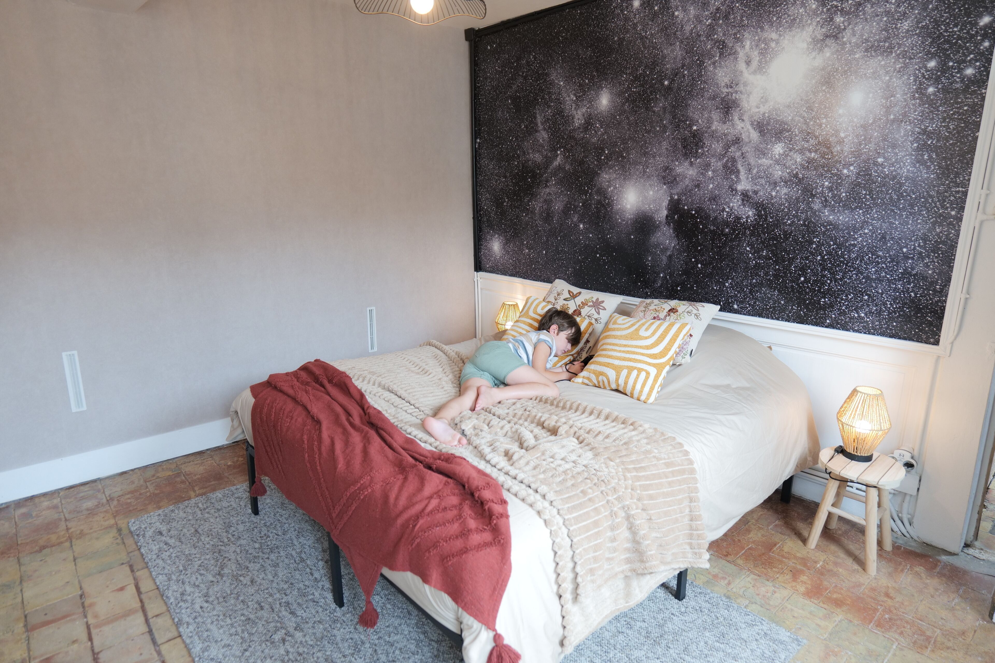 A bedroom with a bed, a nightstand, a lamp, and a wall mural.
