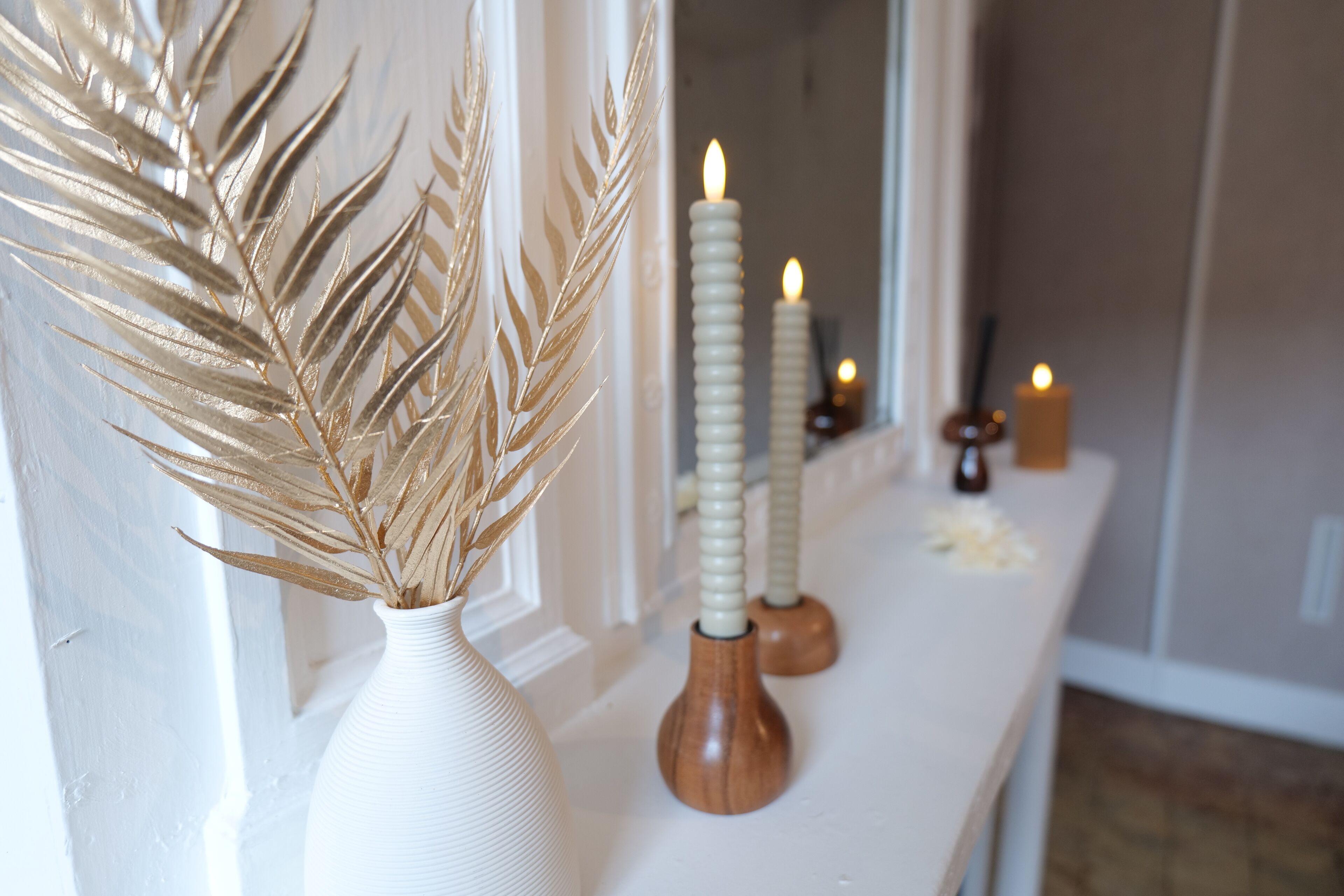Un jarrón blanco con espigas de trigo seco, dos velas con bases de madera y una vela pequeña sobre una mesa en una habitación con una puerta blanca y un espejo.