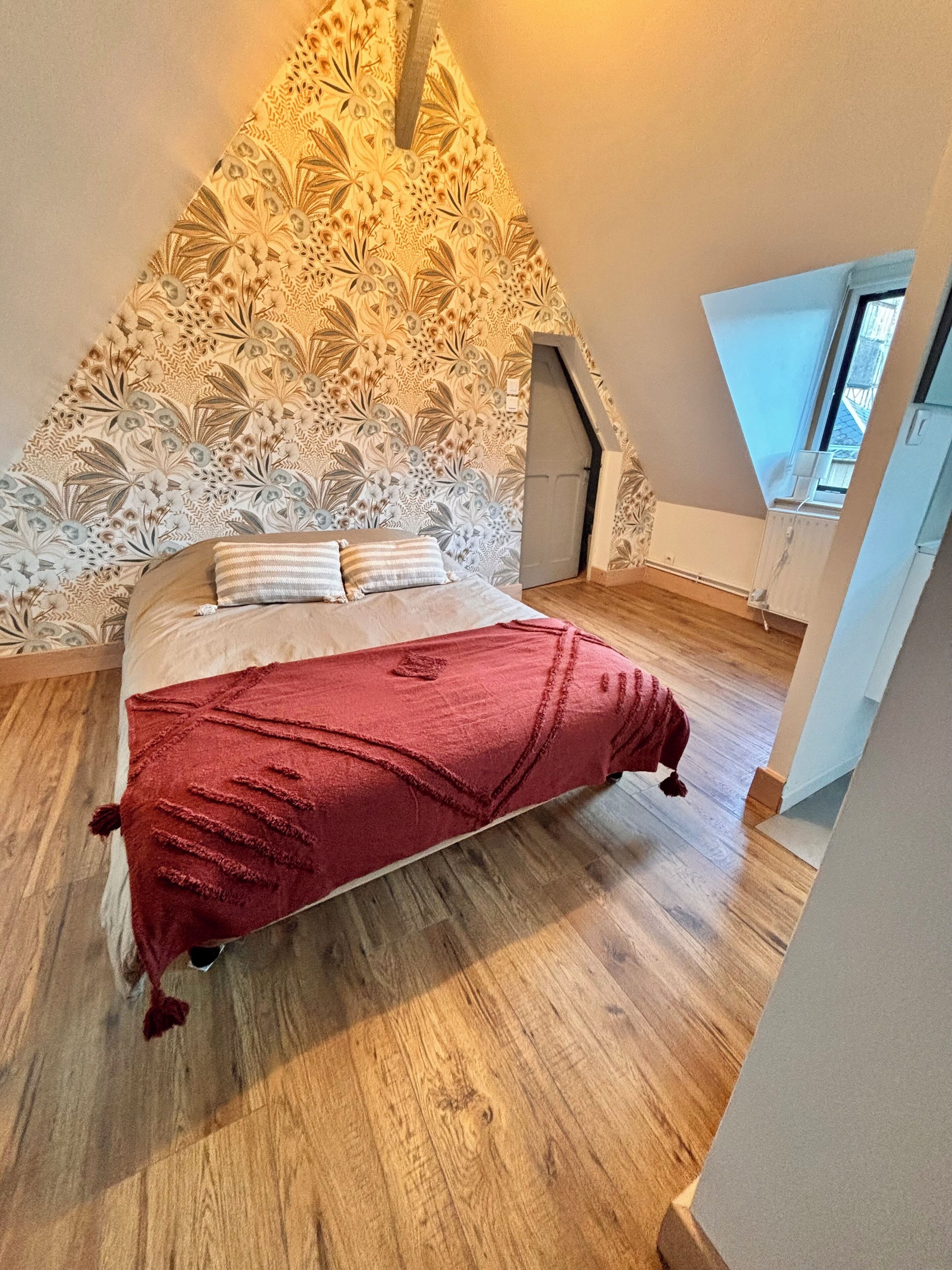 Une chambre avec un plancher en bois, un lit recouvert d’une couverture rouge et un mur décoratif orné de motifs floraux.