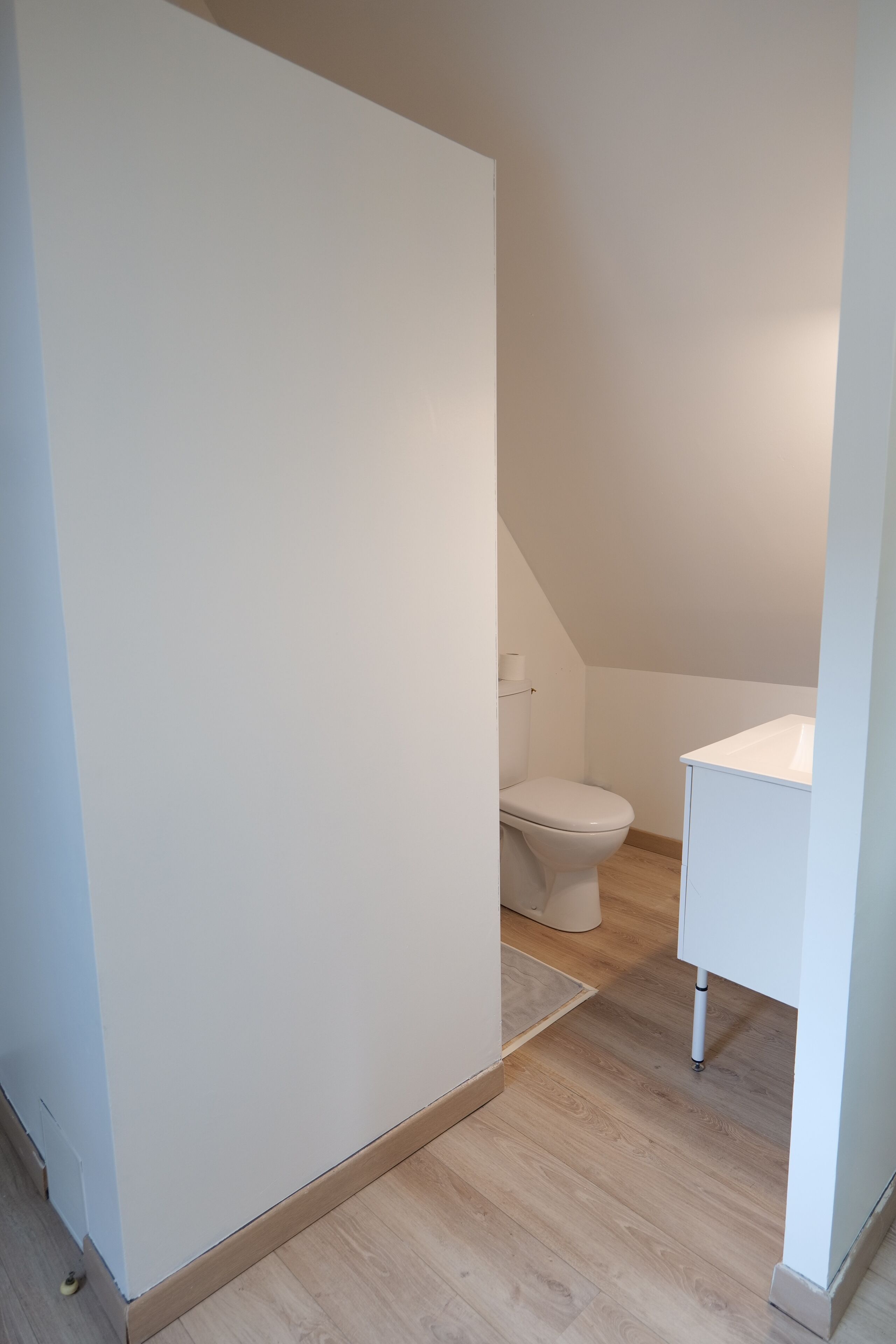 Un baño pequeño y limpio con inodoro y lavamanos blancos, suelo de madera y techo inclinado.
