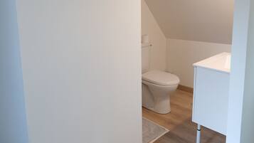 Un baño pequeño y limpio con inodoro y lavamanos blancos, suelo de madera y techo inclinado.