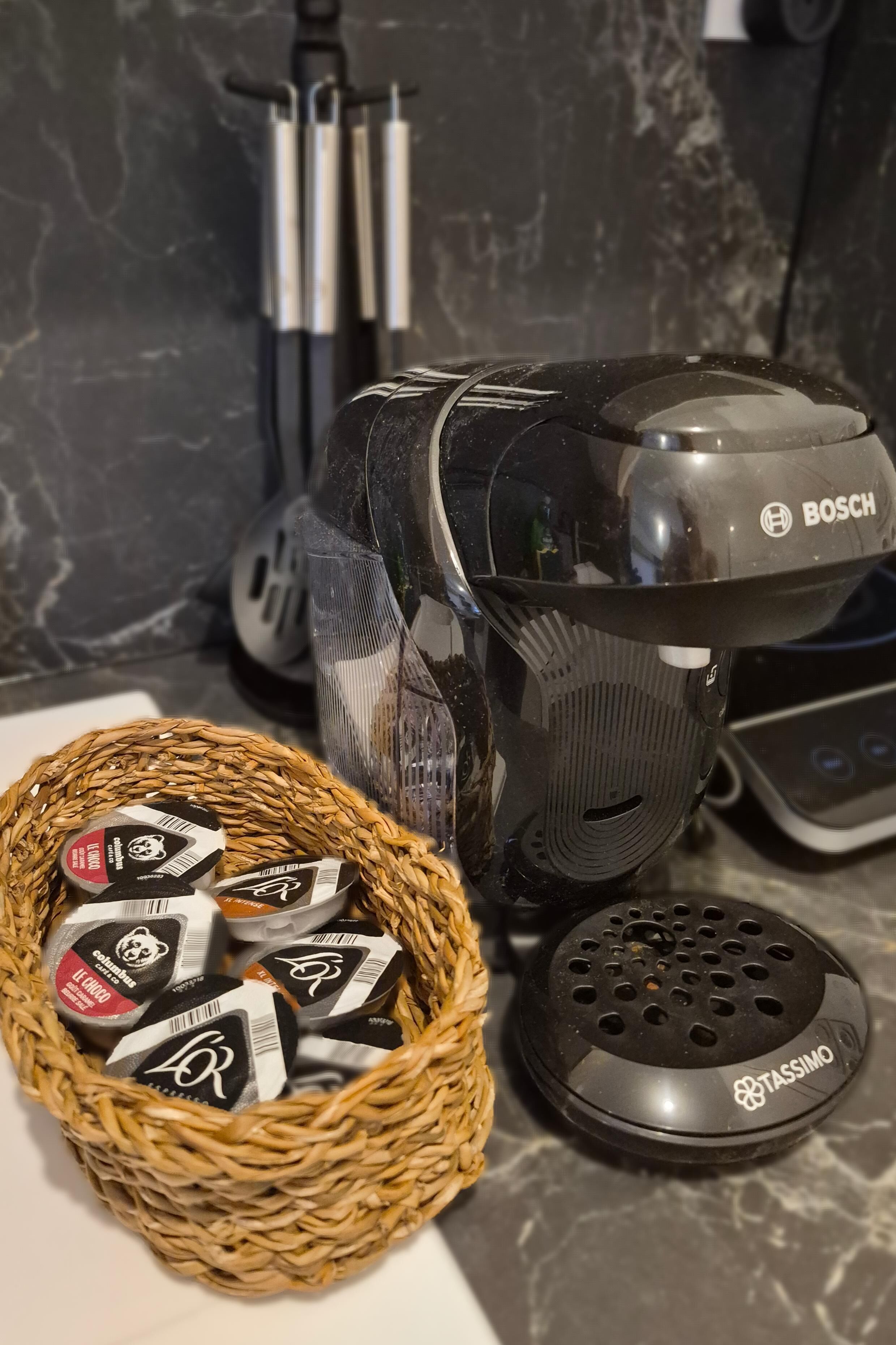 Een koffiezetapparaat, een mand met koffiecups en een Tassimo houder op een aanrecht.