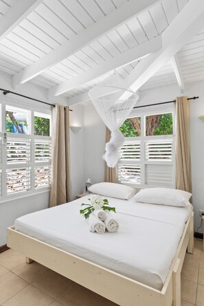Un lit blanc avec des serviettes enroulées et une plante, une fenêtre avec des volets et un plafond avec des poutres en bois.