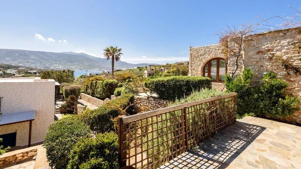 A stone building with a wooden gate, surrounded by well-maintained gardens and a view of the sea and mountains. - Aegean Whisper Home (Paros)