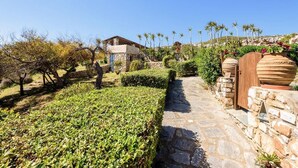 A stone pathway leading to a building with a blue door, surrounded by well-maintained hedges and palm trees. - Aegean Whisper Home (Paros)