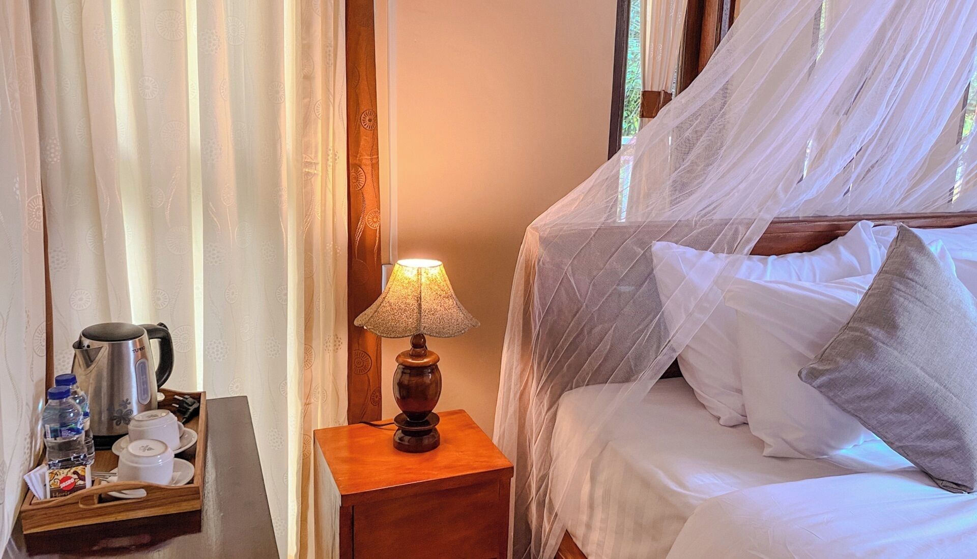 Habitación de hotel con una cama cubierta de sábanas blancas y dosel, una mesita de noche con lámpara, y una bandeja con tetera, tazas y botella de agua.