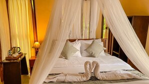 Una cama con dosel y mosquitero, una escultura de toalla, una mesita de noche con lámpara y vistas al exterior por la ventana.