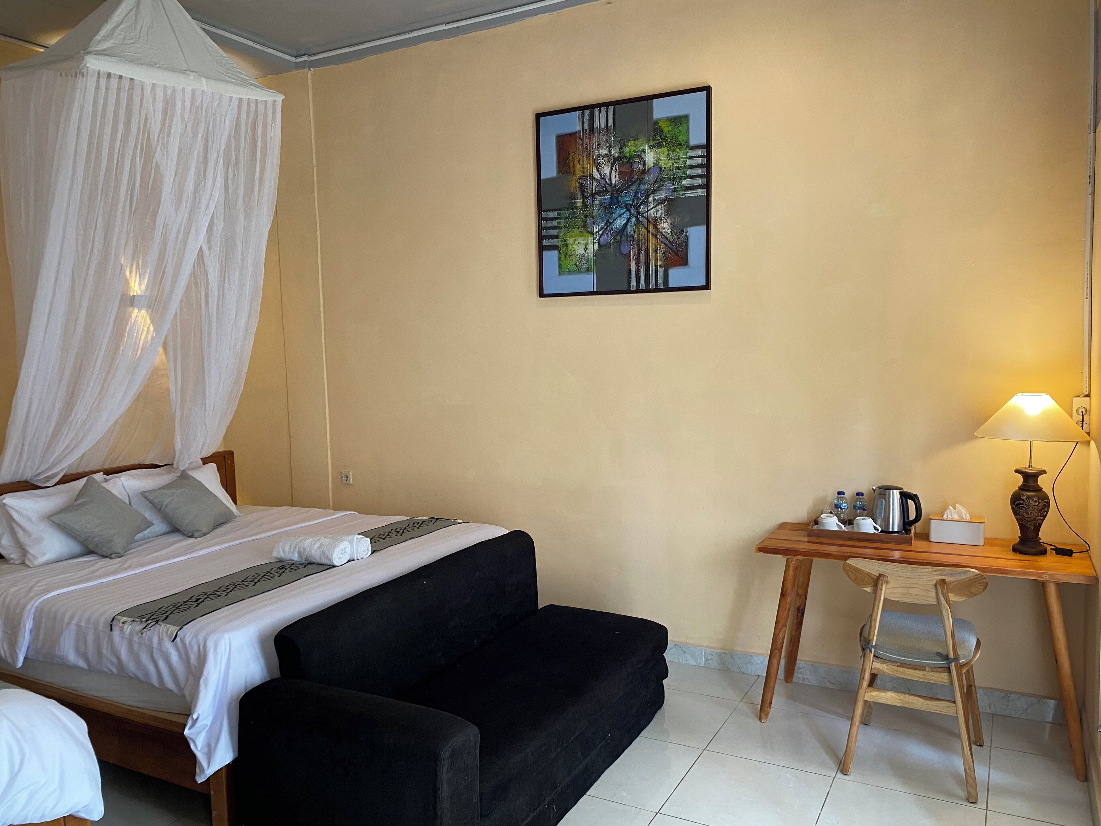 A hotel room with a bed, a black sofa, a desk with a chair, a table lamp, and a painting on the wall.