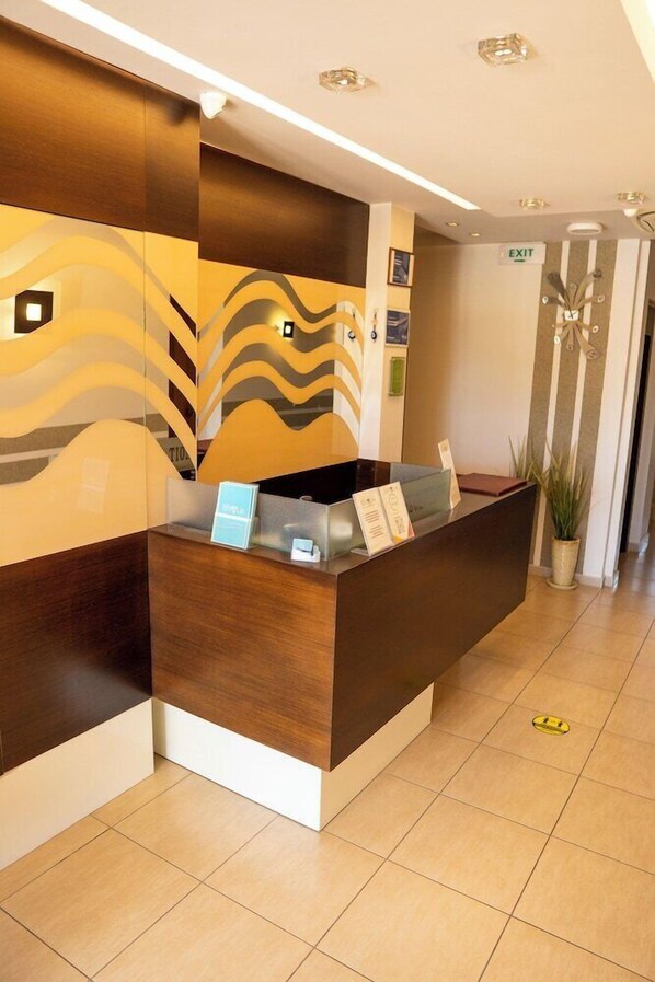 A reception desk with a modern design, a sign indicating an exit, and a potted plant beside it. - Epavlis Hotel (Katerini)