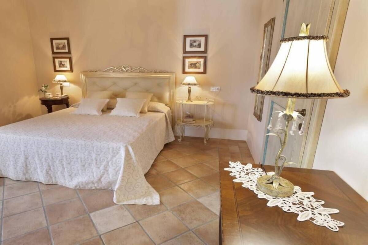 A bedroom with a large bed, two bedside tables with lamps, a decorative lamp on a side table, and framed pictures on the wall.