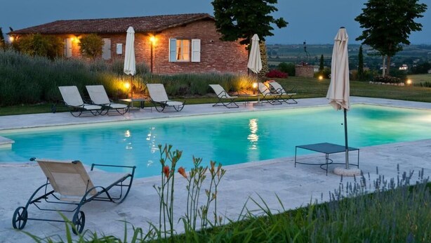 A pool area with lounge chairs and umbrellas, a brick building with lights, and a view of the countryside. - Poggio al Vento Casa Vacanze (Foiano della Chiana)
