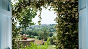 A view through an open door framed by lush greenery and flowers, leading to a scenic landscape with trees and a distant view of a city skyline. - Poggio al Vento Casa Vacanze (Foiano della Chiana)