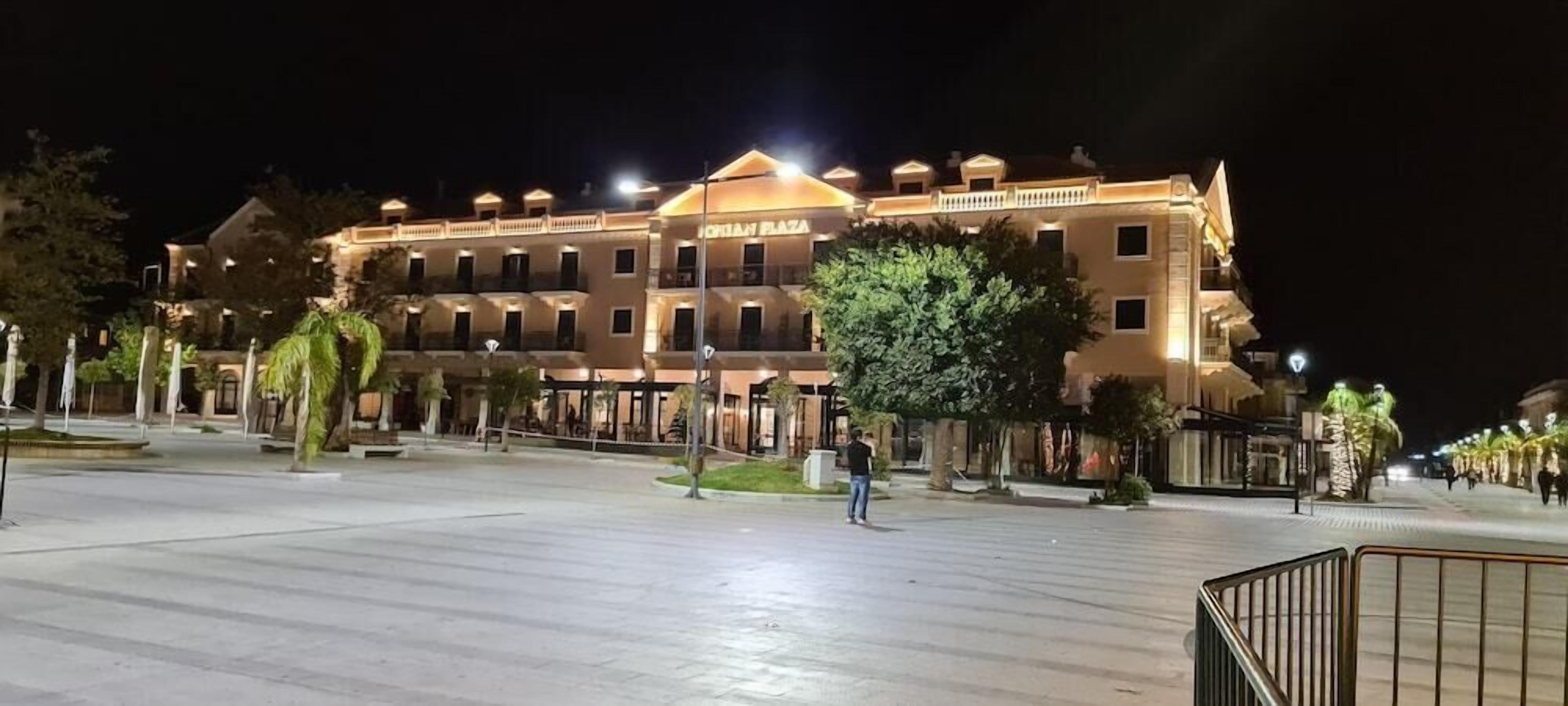 A large, well-lit hotel with multiple floors and balconies, surrounded by trees and a paved plaza.